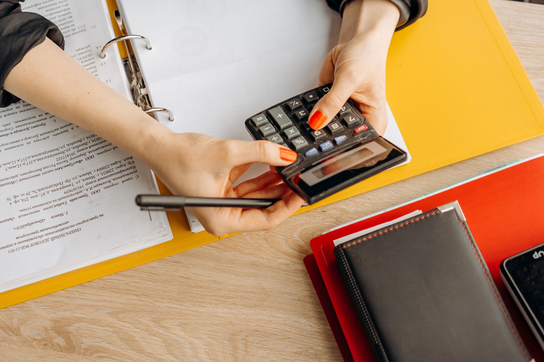 Mains tenant une calculatrice et un stylo au-dessus d'un classeur jaune rempli de documents, sur un bureau en bois