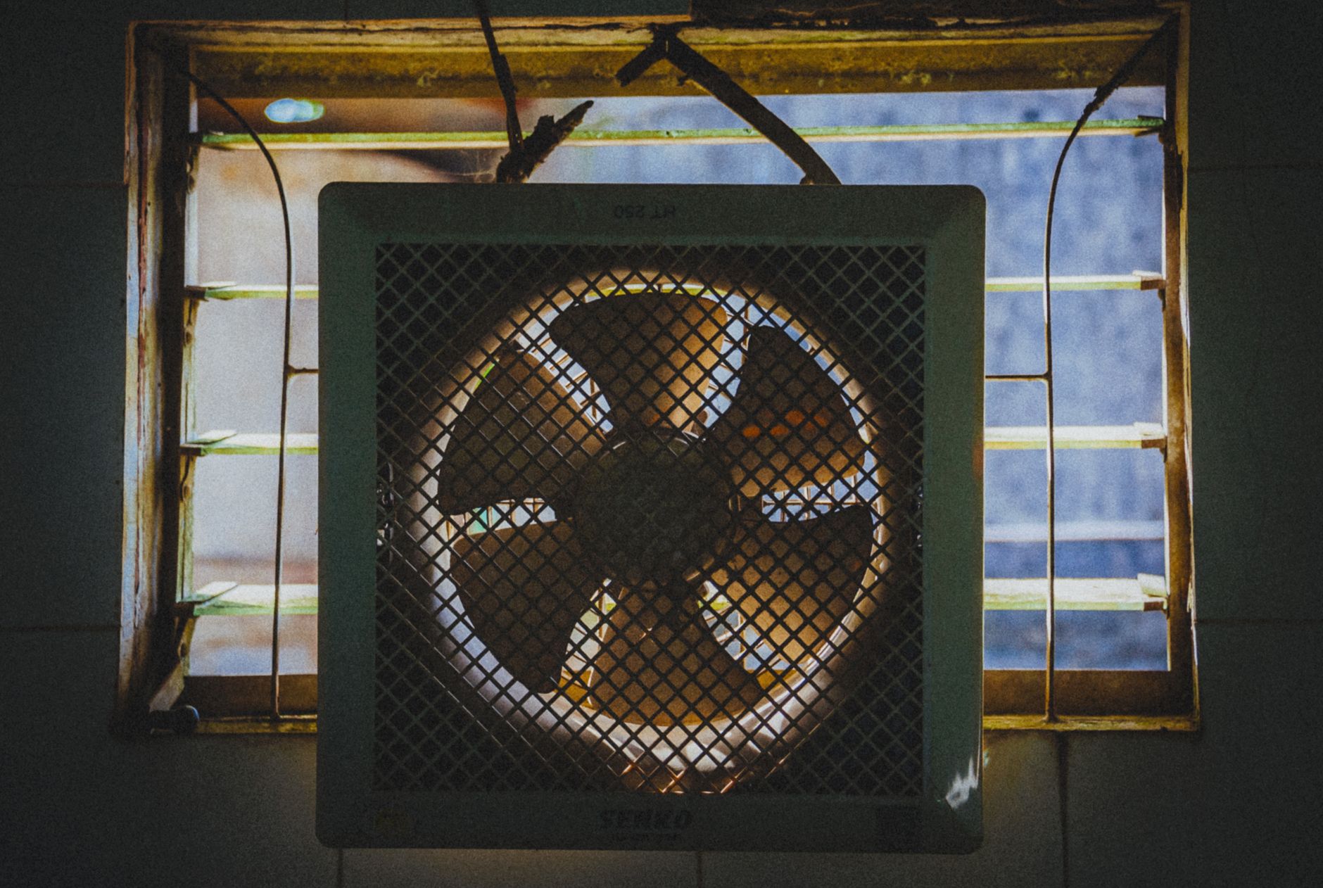 Ancien ventilateur extracteur d'air fixé dans l'encadrement d'une fenêtre à lames, en contre-jour